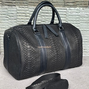 以下が含まれることがあります： 黒色のダッフルバッグ。ヘビ革模様と黒革のハンドルが付いています。バッグには上部にジッパー開閉部があり、取り外し可能なショルダーストラップが付いています。グレーの表面に置かれています。