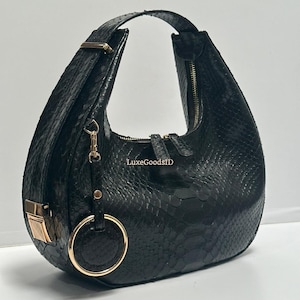 May include: Black handbag with a textured snakeskin pattern. The bag features a curved design, a top handle, and a gold-toned zipper and hardware. A gold ring detail hangs from the front.