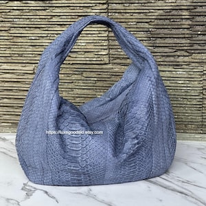 May include: A large, soft, blue handbag with a textured, scale-like pattern. The bag has a single, wide handle and a rounded shape. The background is a textured, stone-like wall. The bag's surface has a reptile-like texture.