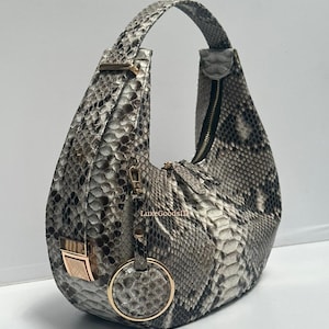 May include: A gray and white snakeskin-patterned handbag with a curved shape and a single shoulder strap. The bag features gold-colored hardware, including a zipper, clasp, and a circular charm. The text "LuxeGoodsIn" is visible on the bag.