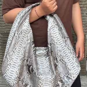 May include: A large, gray and white snakeskin-patterned handbag with a wide shoulder strap. The bag is held by a person wearing a brown t-shirt. The bag's design resembles real snakeskin.