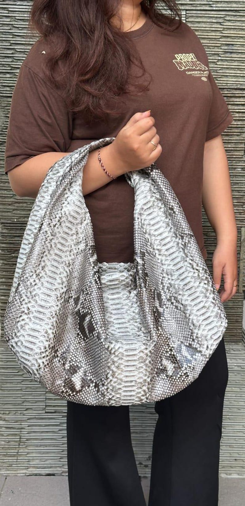 May include: A large, gray and white snakeskin-patterned handbag with a wide shoulder strap. The bag is held by a person wearing a brown t-shirt. The bag's design resembles real snakeskin.