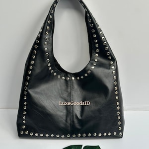 May include: Black hobo bag with silver-colored stud accents along the edges. The bag has a single shoulder strap and a smooth, matte finish. The text "LuxeGoodsID" is visible on the front of the bag.
