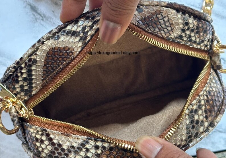 Peut inclure: Sac &agrave; main rond avec un motif peau de serpent marron, beige et noir. Il est dot&eacute; d'une fermeture &eacute;clair dor&eacute;e et d'une bandouli&egrave;re cha&icirc;ne dor&eacute;e. L'int&eacute;rieur est de couleur marron clair. Le sac est ouvert.