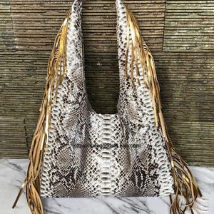 Puede incluir: Bolso de mano espacioso con estampado de piel de serpiente en blanco, gris y negro. El bolso tiene flecos dorados en los laterales y un asa ancha y curvada. Un accesorio de moda.