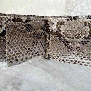 Puede incluir: Cartera bi-fold abierta hecha de piel de serpiente. La cartera est&aacute; abierta, revelando m&uacute;ltiples ranuras para tarjetas y un compartimento para billetes. El patr&oacute;n de piel de serpiente presenta tonos de marr&oacute;n, gris y blanco. La cartera se muestra sobre una superficie blanca y esponjosa.