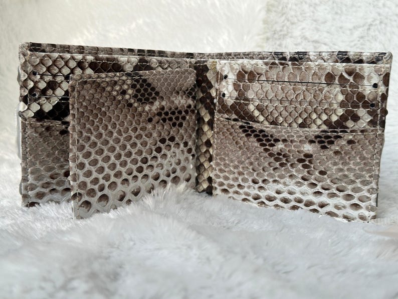 Puede incluir: Cartera bi-fold abierta hecha de piel de serpiente. La cartera est&aacute; abierta, revelando m&uacute;ltiples ranuras para tarjetas y un compartimento para billetes. El patr&oacute;n de piel de serpiente presenta tonos de marr&oacute;n, gris y blanco. La cartera se muestra sobre una superficie blanca y esponjosa.