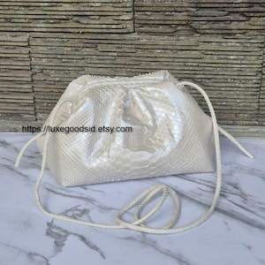 Op de afbeelding: Een witte, getextureerde handtas met een trekkoordsluiting en een lang, wit koord. De tas heeft een parelmoerglans en een reptielachtig patroon. De Etsy shop URL is zichtbaar.