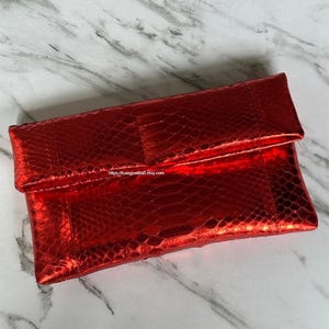 May include: A red clutch with a snakeskin pattern. The rectangular bag has a fold-over flap and a shiny, textured surface. The Etsy shop URL is visible on the bottom of the image.