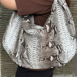 May include: A large, rounded shoulder bag with a snakeskin pattern in shades of white, gray, and brown. The bag is worn over the shoulder, and the strap is wide. The bag appears to be made of a textured material.