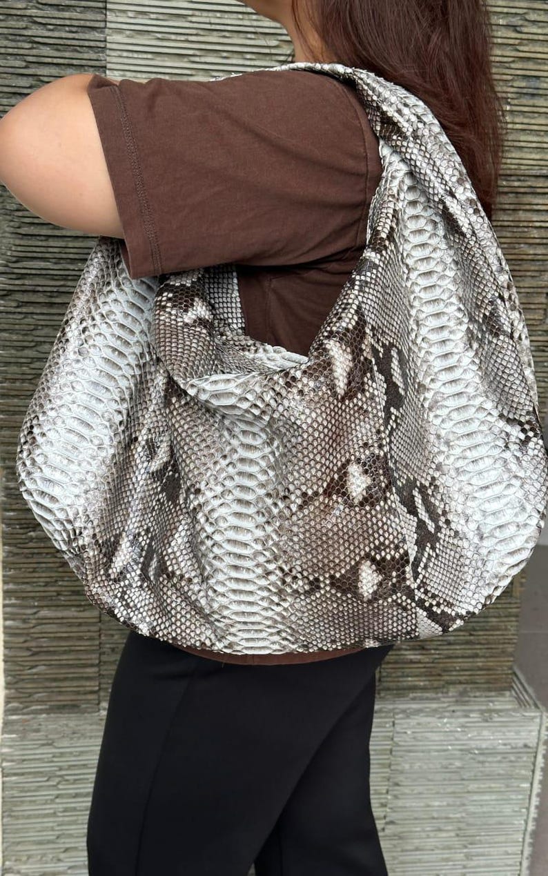 May include: A large, rounded shoulder bag with a snakeskin pattern in shades of white, gray, and brown. The bag is worn over the shoulder, and the strap is wide. The bag appears to be made of a textured material.