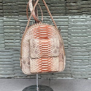 May include: A small, round backpack made of snakeskin in shades of tan, brown, and orange. The bag has a front pocket and a top handle, and is displayed on a silver stand. The text "LuxeGoodsShop" is visible on the front pocket.