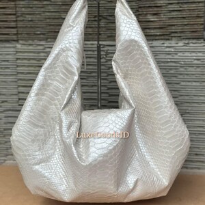 Puede incluir: Un bolso blanco perlado con un estampado de piel de serpiente. El bolso tiene un diseño grande y abierto con un solo asa. El bolso se exhibe sobre un soporte metálico contra un fondo texturizado. El texto "LuxeGoodsID" es visible.