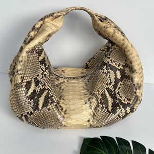 May include: A snakeskin hobo bag in beige and brown tones, with a curved handle. The bag displays a natural snakeskin pattern with dark brown and beige scales. It is set against a white backdrop.