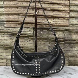 Puede incluir: Bolso de hombro de cuero negro con detalles de tachuelas y ojales plateados. El bolso en forma de media luna tiene cierre de cremallera y una correa larga. La URL de la tienda Etsy es visible.
