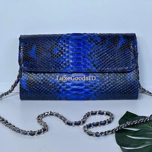 May include: A blue and black snakeskin-patterned clutch with a silver chain strap. The clutch has a flap closure and the text "LuxeGoodsID" is visible on the front. The bag is on a white surface.