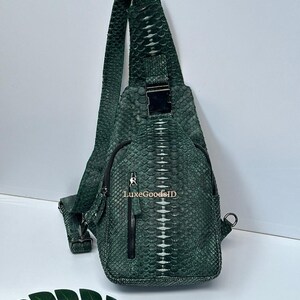 May include: A green, textured sling bag with a single shoulder strap. The bag features a front zipper pocket, a silver-colored metal buckle, and a small metal ring. The text "LuxeGoodsID" is visible on the front of the bag.