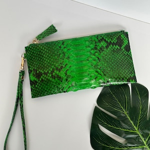 May include: A vibrant green clutch bag with a snakeskin design and a gold-coloured zip. The bag features a matching wrist strap and a gold zip pull. The background is white.