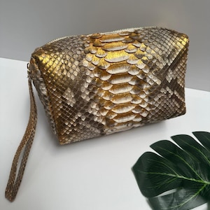 Könnte beinhalten: Eine rechteckige Clutch aus gold- und braunfarbenem Schlangenleder mit einer Handschlaufe. Die Clutch hat einen Reißverschluss und eine goldfarbene Mitte. Das Schlangenmuster ist auf der Außenseite der Tasche sichtbar.