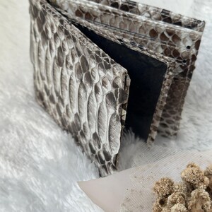 Puede incluir: Una cartera plegable con un patr&oacute;n de piel de serpiente en tonos grises y marrones. La cartera est&aacute; abierta, revelando un interior negro. La superficie texturizada de la cartera es visible, mostrando las escamas. La cartera est&aacute; sobre una superficie blanca y esponjosa.