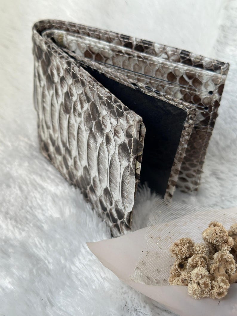 Puede incluir: Una cartera plegable con un patr&oacute;n de piel de serpiente en tonos grises y marrones. La cartera est&aacute; abierta, revelando un interior negro. La superficie texturizada de la cartera es visible, mostrando las escamas. La cartera est&aacute; sobre una superficie blanca y esponjosa.
