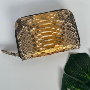 May include: A small, zippered wallet with a snakeskin pattern in shades of gold, brown, and tan. The wallet has a black zipper and a matching pull tab. The overall design is compact and stylish.