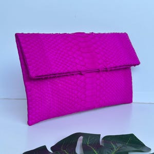 Puede incluir: Un bolso de mano fucsia brillante con un patrón de piel de serpiente texturizado. El bolso tiene forma rectangular con cierre de solapa plegada. El fondo es blanco.