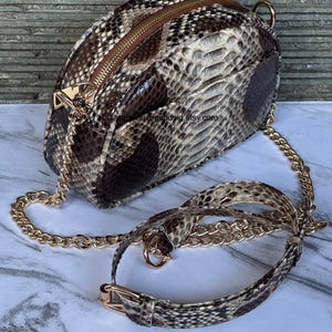 Peut inclure: Sac bandouli&egrave;re &agrave; motif peau de serpent avec une cha&icirc;ne dor&eacute;e et une fermeture &eacute;clair. Le sac pr&eacute;sente un motif marron, beige et noir. La bandouli&egrave;re comprend une section &agrave; motif peau de serpent avec une boucle dor&eacute;e.