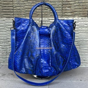 Könnte beinhalten: Eine leuchtend blaue Handtasche mit Schlangenmuster. Die Tasche hat einen oberen Griff und einen abnehmbaren Schultergurt. Der Text "LuxeGoodsID" ist auf der Tasche sichtbar. Die Tasche wird auf einem silbernen Ständer präsentiert.