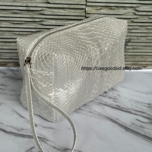 Könnte beinhalten: Eine perlweiße Kosmetiktasche mit strukturiertem Schlangenmuster. Die rechteckige Tasche hat einen silbernen Reißverschluss und eine passende Handschlaufe. Die Etsy-Shop-URL ist sichtbar.