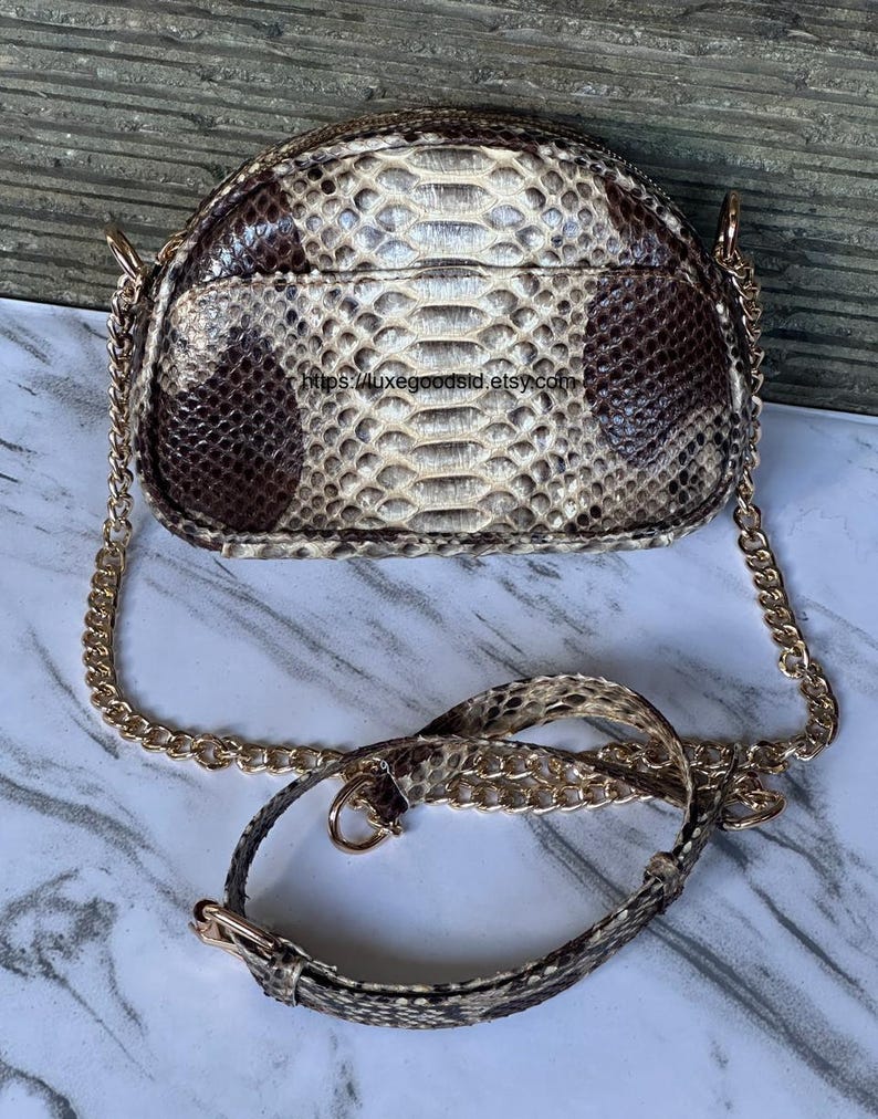 Peut inclure: Un petit sac &agrave; main rond avec un motif peau de serpent dans les tons marron, beige et cr&egrave;me. Le sac est dot&eacute; d'une bandouli&egrave;re en cha&icirc;ne dor&eacute;e et d'une bandouli&egrave;re assortie en peau de serpent. Le sac a une fermeture &eacute;clair.
