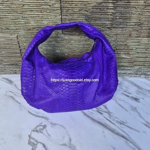 Puede incluir: Un vibrante bolso morado con un patrón texturizado similar a la piel de serpiente. El bolso tiene una sola correa para el hombro y una forma curva. La URL de la tienda Etsy es visible en el bolso. El bolso está sobre un fondo de piedra y mármol.
