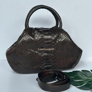 Op de afbeelding: Een bruine leren handtas met een handvat aan de bovenkant en een afneembare schouderriem. De tas heeft een textuur slangenleerpatroon en de tekst "LuxeGoodsID" is zichtbaar aan de voorkant.