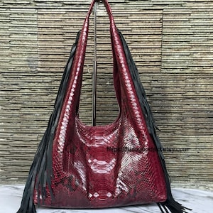 May include: A burgundy leather shoulder bag with a snakeskin pattern and long black fringe. The bag has long handles and is displayed on a metal stand. The bag's texture and color are prominent.