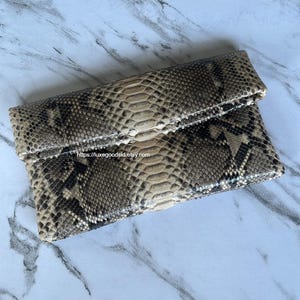 Può includere: Pochette rettangolare con motivo a pelle di serpente nei toni del marrone, beige e nero. La borsa ha una patta. L'URL del negozio Etsy è visibile sull'immagine. La borsa è probabilmente realizzata in pelle o materiale simile.