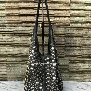 Puede incluir: Un bolso de hombro negro estilo hobo que presenta una densa disposición de tachuelas metálicas circulares de tono plateado en la superficie frontal. El bolso tiene una sola correa integrada y el texto https://luxegoodsid.etsy.com impreso al frente.