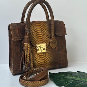 Puede incluir: Un bolso de mano de piel de serpiente marrón y dorada con asa superior y bandolera desmontable. El bolso presenta un cierre dorado y una borla decorativa. El bolso mide aproximadamente 25 cm de ancho y 20 cm de alto.