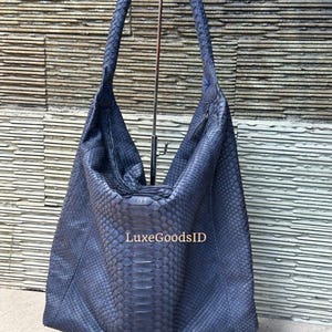 May include: A dark blue leather handbag with a textured snakeskin pattern. The bag has a braided handle and a large, open design. The text "LuxeGoodsID" is visible on the bag.