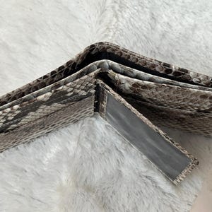Puede incluir: Cartera de dos pliegues abierta con un patr&oacute;n de piel de serpiente en tonos grises, marrones y blancos. La cartera est&aacute; hecha de un material texturizado y tiene m&uacute;ltiples compartimentos para tarjetas y efectivo. La cartera est&aacute; abierta, mostrando su interior.