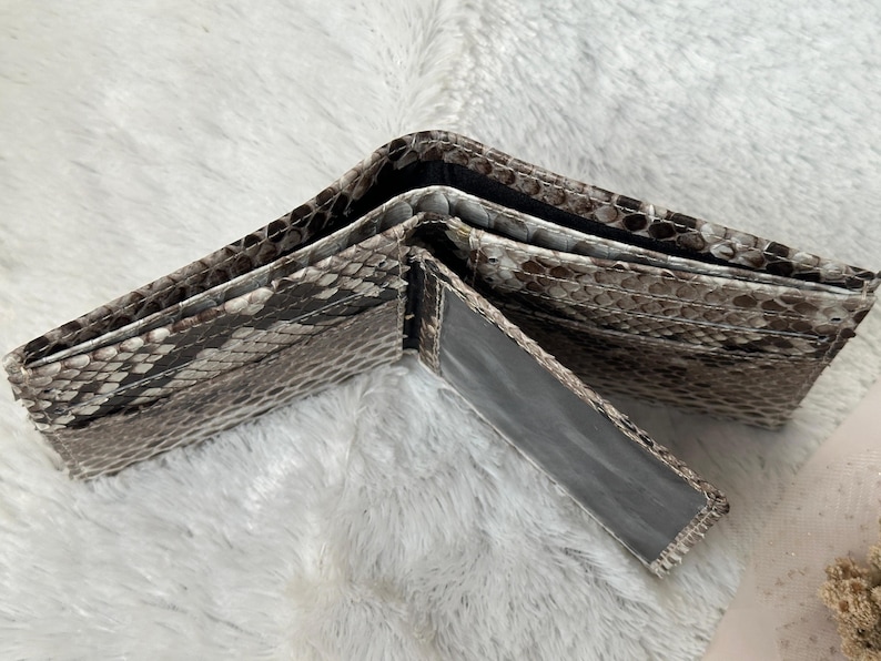 Puede incluir: Cartera de dos pliegues abierta con un patr&oacute;n de piel de serpiente en tonos grises, marrones y blancos. La cartera est&aacute; hecha de un material texturizado y tiene m&uacute;ltiples compartimentos para tarjetas y efectivo. La cartera est&aacute; abierta, mostrando su interior.