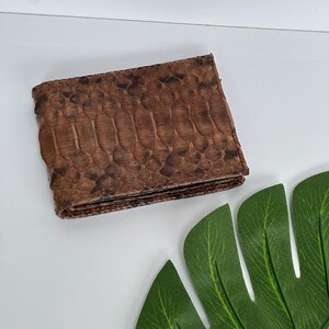 May include: A brown bi-fold wallet with a textured snakeskin pattern. The wallet is made of a dark brown material with lighter brown and black accents. The wallet is rectangular and appears to be closed.
