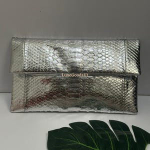 Pode incluir: Uma clutch prateada com padrão de pele de cobra. A bolsa retangular tem uma aba e é feita de um material brilhante e texturizado. O texto "LuxeGoodsID" é visível na bolsa.