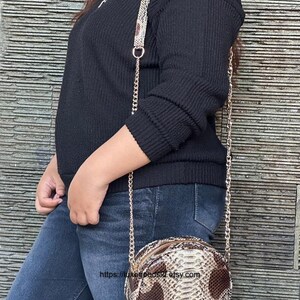 Peut inclure: Un petit sac &agrave; bandouli&egrave;re ovale avec un motif peau de serpent dans des tons marron et beige. Le sac a une bandouli&egrave;re en cha&icirc;ne dor&eacute;e et une fermeture &eacute;clair. Le sac est port&eacute; avec un pull noir et un jean bleu.