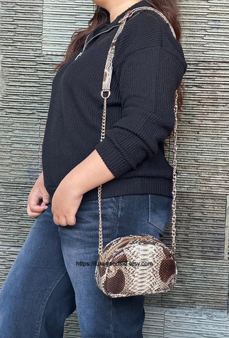 Peut inclure: Un petit sac &agrave; bandouli&egrave;re ovale avec un motif peau de serpent dans des tons marron et beige. Le sac a une bandouli&egrave;re en cha&icirc;ne dor&eacute;e et une fermeture &eacute;clair. Le sac est port&eacute; avec un pull noir et un jean bleu.