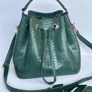 May include: A green bucket bag with a snakeskin pattern. The bag has a drawstring closure, a top handle, and a long, adjustable shoulder strap. Gold-colored hardware accents the bag.
