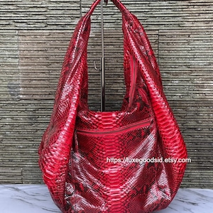 Op de afbeelding: Een grote, rode schoudertas met slangenleertextuur, ritssluiting en lange riem. De tas wordt gepresenteerd op een metalen standaard tegen een getextureerde stenen muur. De Etsy shop URL is zichtbaar.