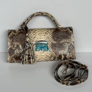 May include: A snakeskin-patterned handbag with a top handle and a detachable shoulder strap. The bag features a rectangular shape with a flap closure and a decorative tassel. The hardware is gold-toned, and the bag has a light blue clasp.