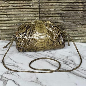 May include: A snakeskin shoulder bag in shades of brown and tan, with a drawstring closure and a long, thin strap. The bag has a rounded shape and a textured surface, with a marble background.