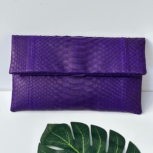 May include: A vibrant purple clutch with a textured snakeskin pattern. The rectangular bag features a fold-over flap and is made of a durable material. The clutch is a stylish accessory for various occasions.