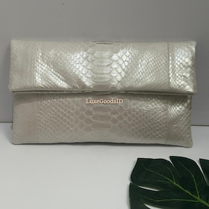 May include: A pearlescent white clutch with a snakeskin pattern. The rectangular bag has a fold-over flap and is made of a textured material. The text "LuxeGoodsID" is visible on the front.
